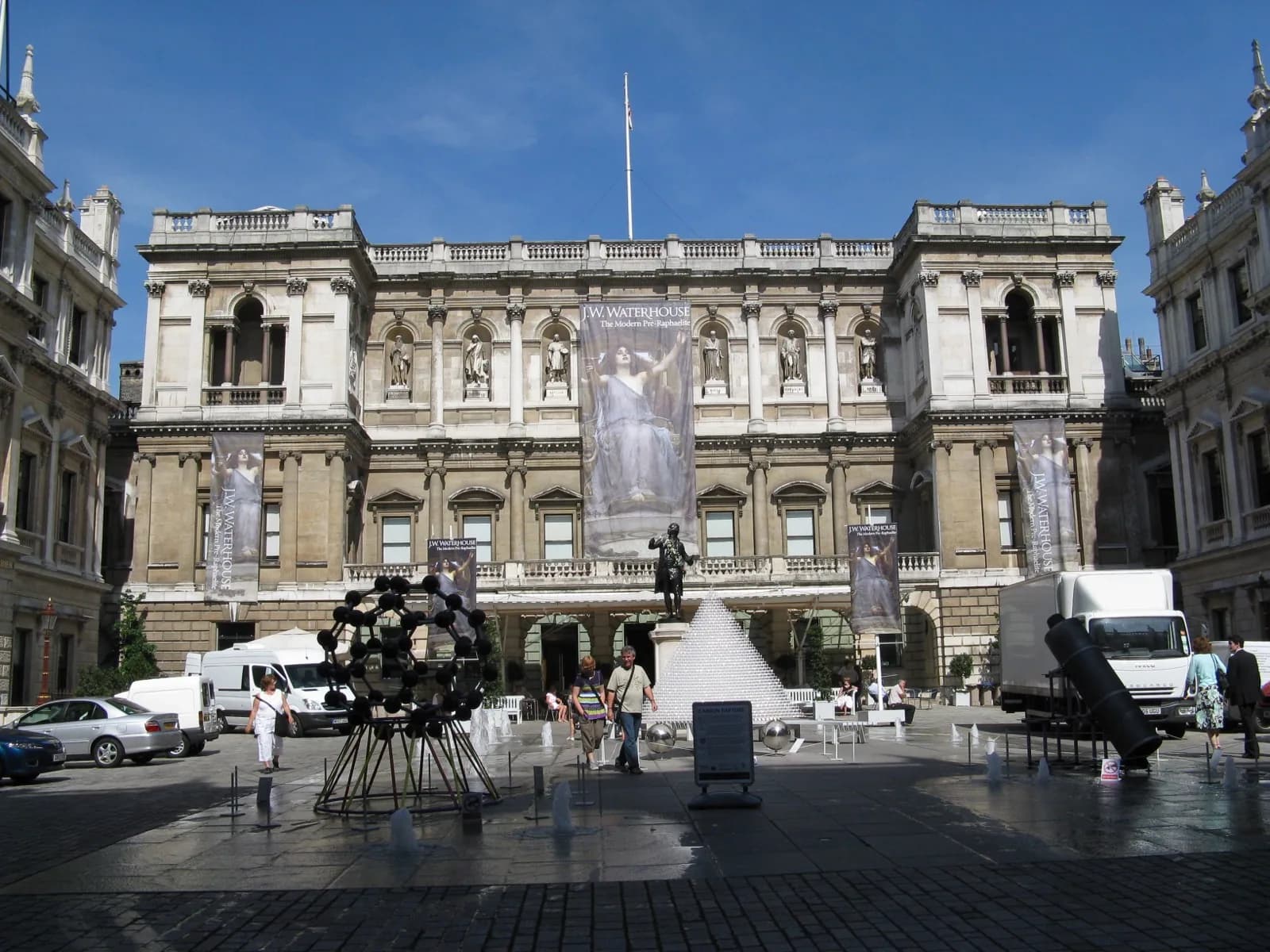 Courtyard view of Burlington House, home of the Royal Academy of Arts in London, showing the grand portico with columns, statues of artists including Leonardo da Vinci, and visitors on a sunny day.