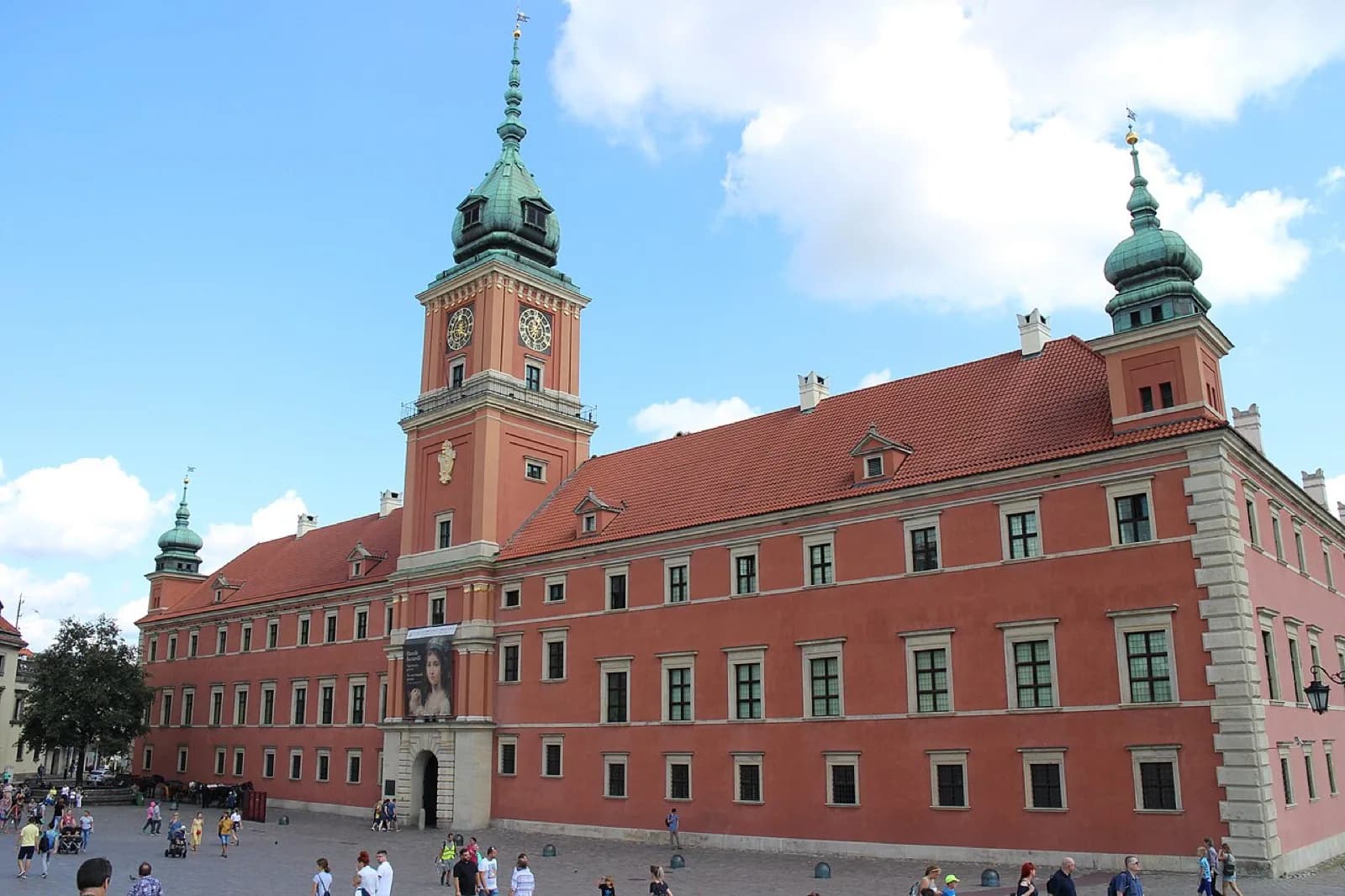 Daytime view of the Royal Castle in Warsaw's red baroque facade with its central clock tower and green domes, from the cobblestone Castle Square with visitors, Poland.