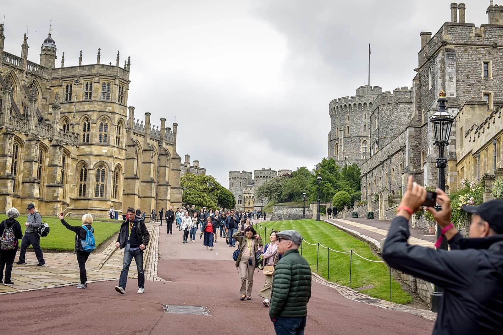Overcast view of Windsor Castle's historic stone facade and towers with visitors walking along the path and lawn in the castle grounds, Windsor, United Kingdom.