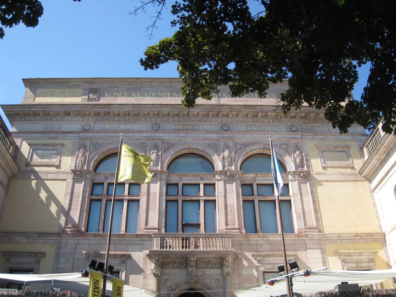 The facade of the Royal Swedish Academy of Fine Arts building in Stockholm, Sweden, a classical stone building with arched windows, statues, and inscriptions under a blue sky.