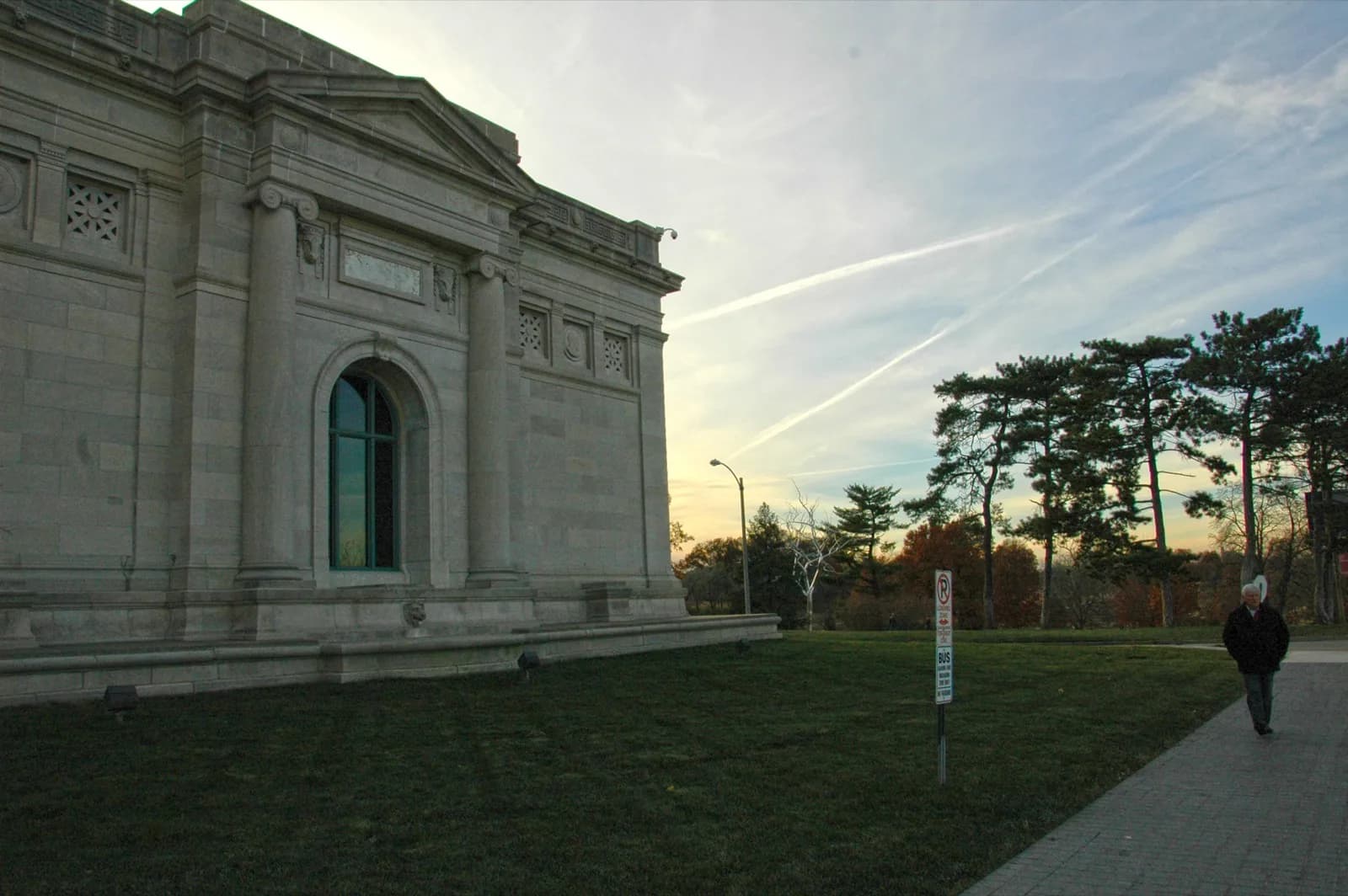 Grand stone facade of the Saint Louis Art Museum's 1904 World's Fair building in Forest Park, featuring classical columns, arched window, and pediment details against a sunset sky with autumn trees.