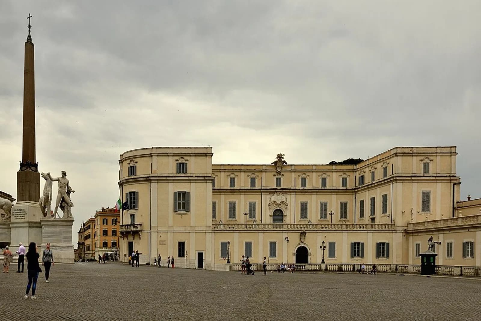 The long, curved neoclassical facade of the Scuderie del Quirinale exhibition hall in Rome's Piazza del Quirinale, a pale yellow building with arched windows and central entrance, viewed across the cobblestone square with the Quirinal obelisk, Dioscuri statues, and visitors.