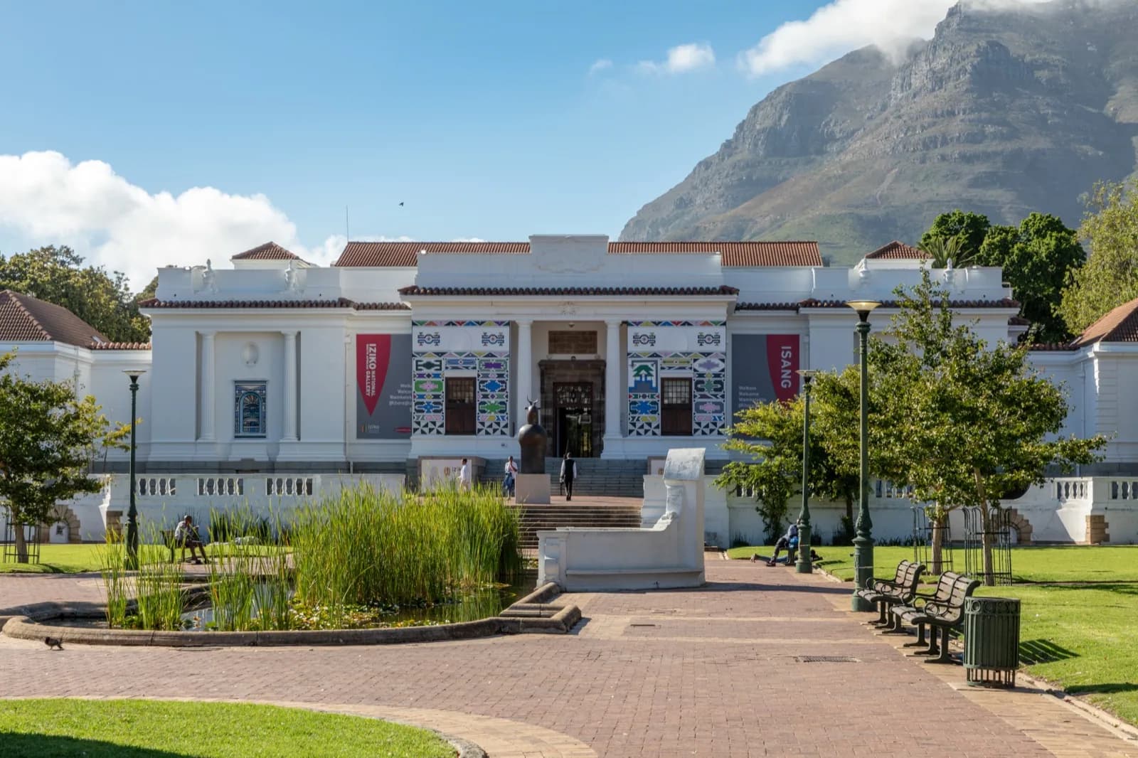 The Iziko South African National Gallery in Cape Town, a white neoclassical building with colorful decorative panels and banners, viewed from the gardens with Table Mountain in the background on a sunny day.