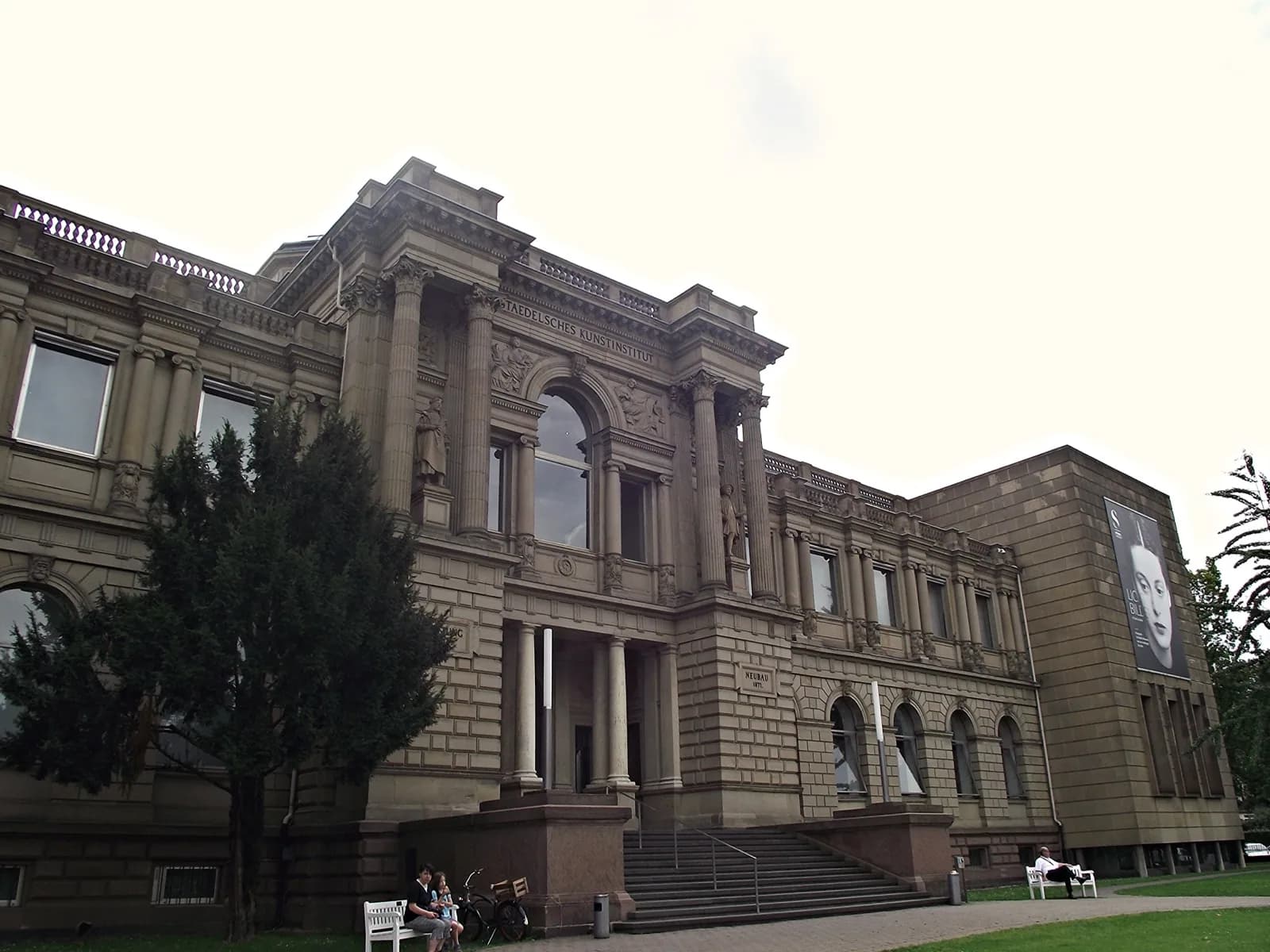 View of the grand stone facade and entrance stairs of the Städel Museum in Frankfurt, Germany, with its classical columns, sculptures, and adjacent modern wing, on an overcast day.