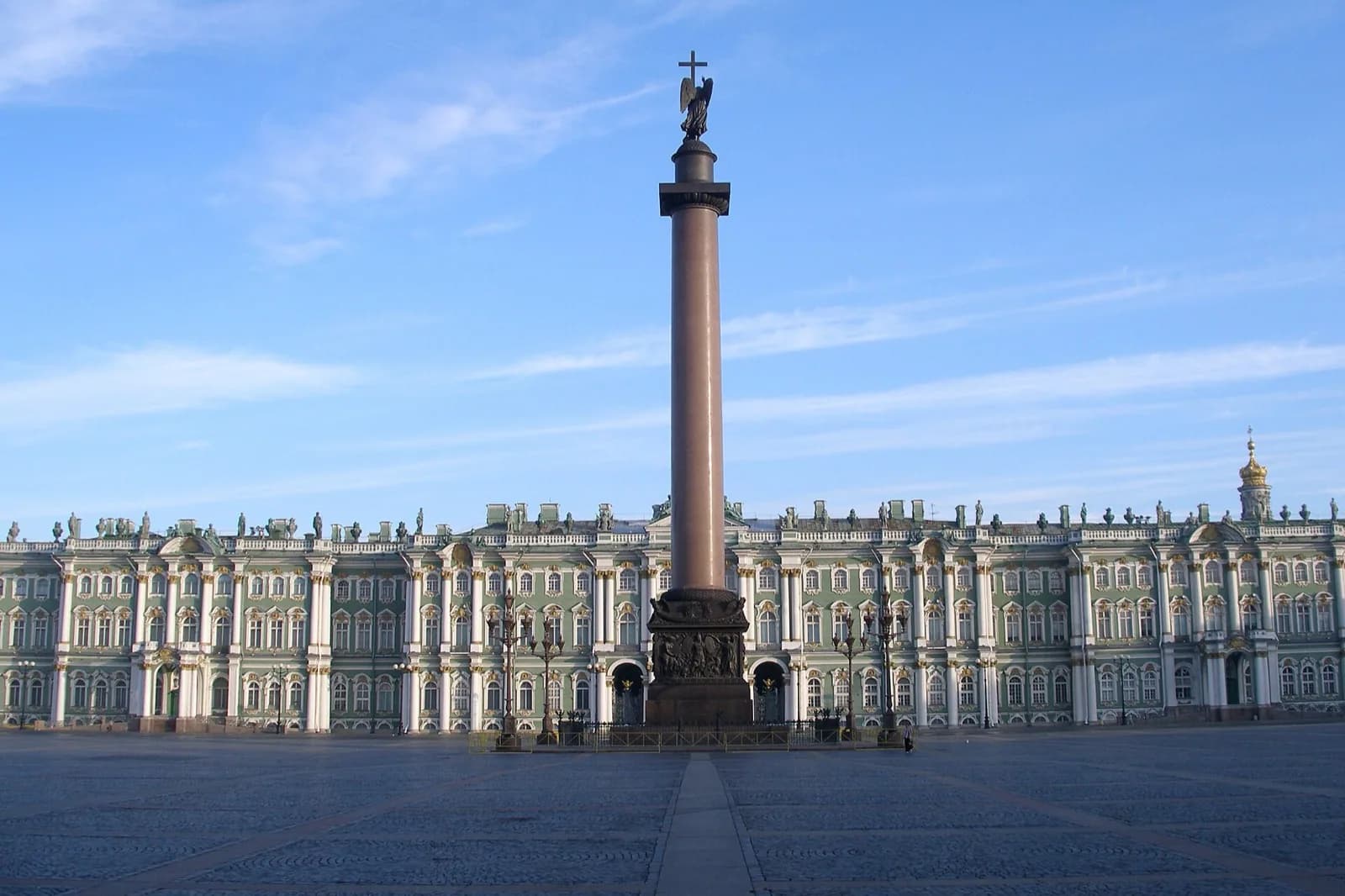 The grand green-and-white baroque facade of the Winter Palace (State Hermitage Museum) along Palace Square in Saint Petersburg, Russia, with the tall Alexander Column topped by a winged angel statue centered in the vast cobblestone square under a bright blue sky.