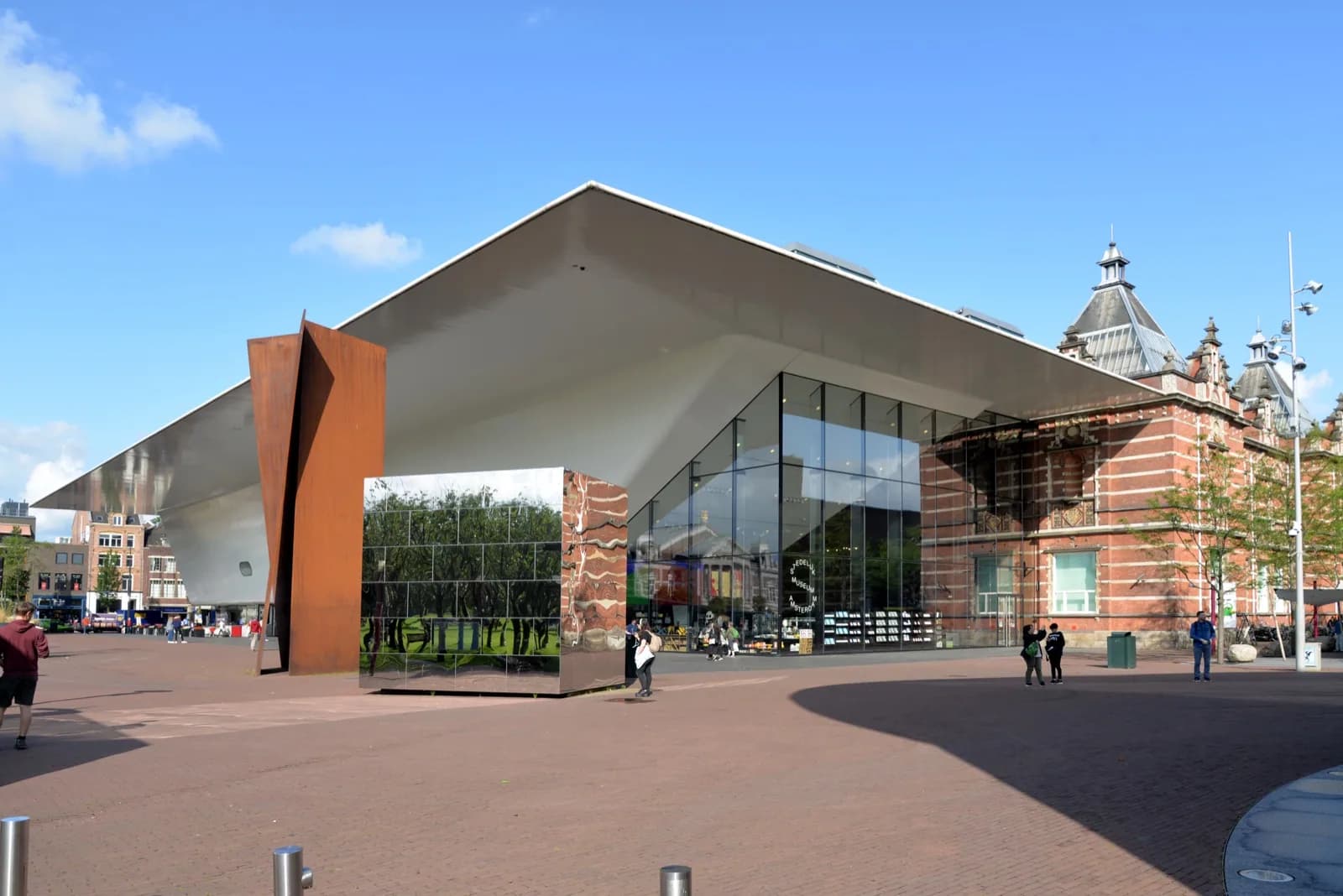 The modern Benthem Crouwel Wing of the Stedelijk Museum in Amsterdam, a striking white and glass structure with a large reflective red panel, in front of the historic red brick building on Museumplein.