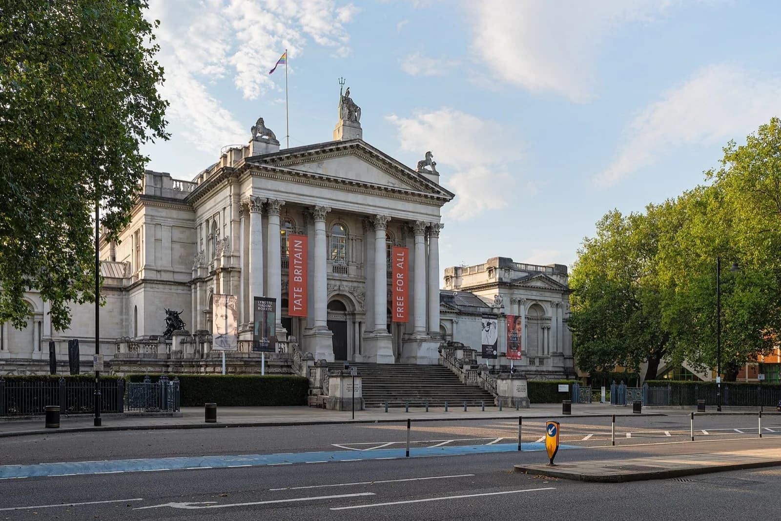 The neoclassical portico of Tate Britain on the bank of the Thames in London, with its Ionic columns, pediment sculptures, and red banners, set in a plaza with trees under a partly cloudy sky.