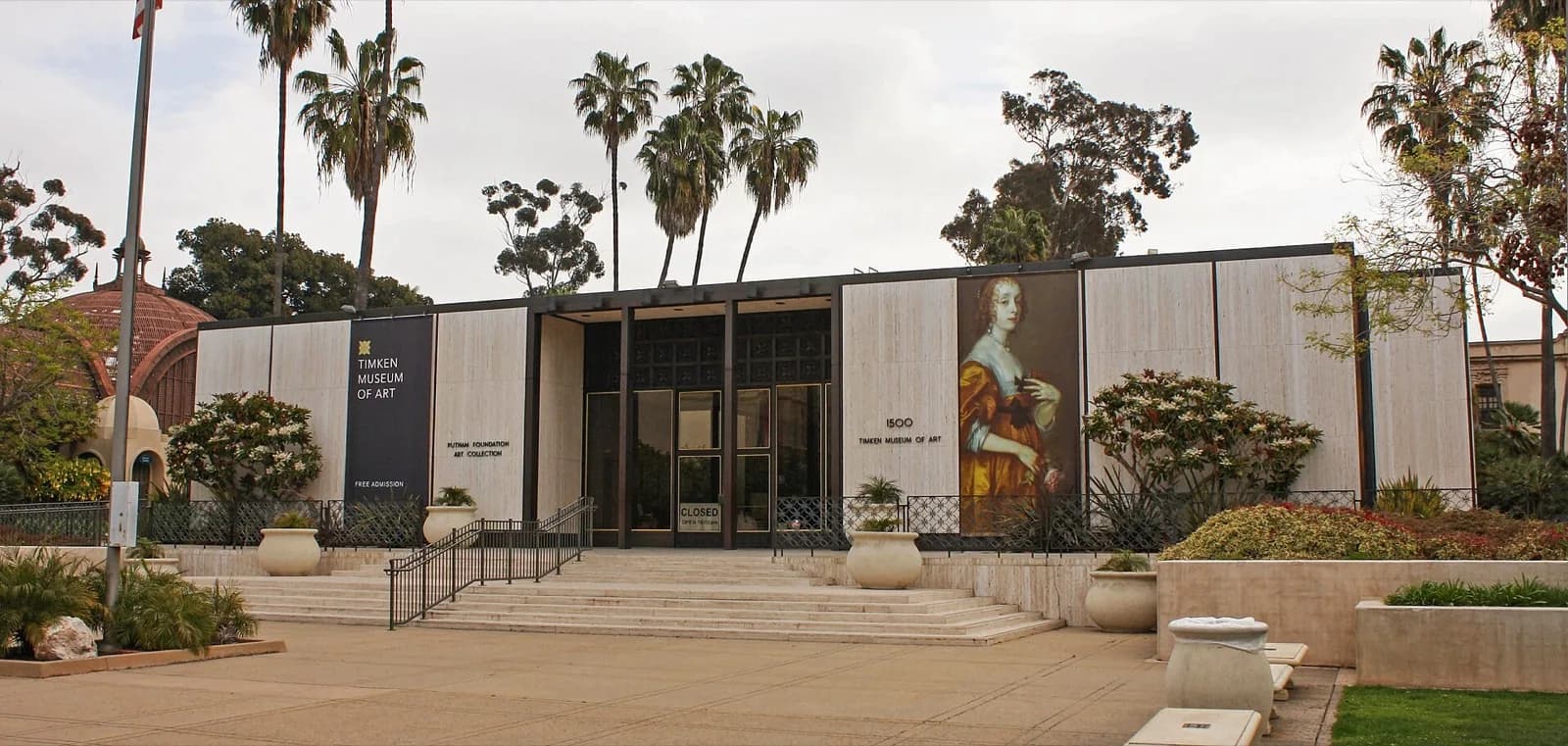 The modern facade of the Timken Museum of Art in Balboa Park, San Diego, California, with light-colored wood paneling, a prominent reproduction of a historical portrait on the right, central glass entrance doors, potted plants, flower beds, and tall palm trees under a bright partly cloudy sky.