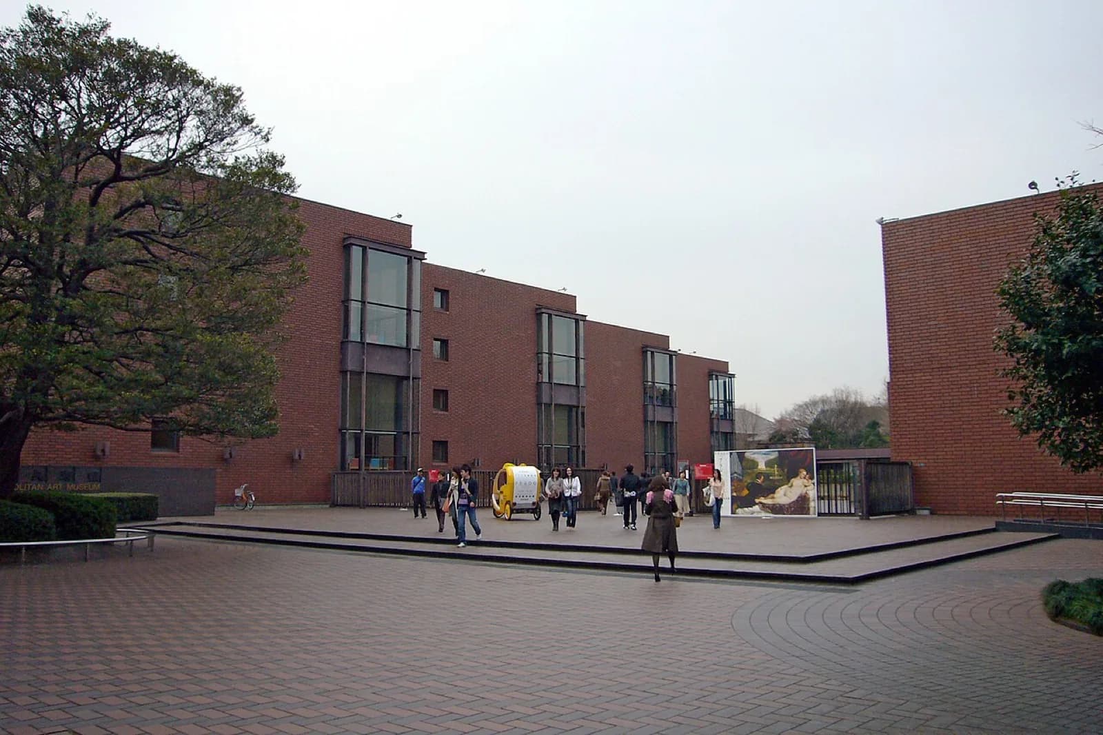 The modern red-brick exterior of the Tokyo Metropolitan Art Museum in Ueno Park, Tokyo, featuring large vertical glass windows, a spacious paved forecourt with visitors, a large reproduction of a classical painting on display, and surrounding trees under an overcast sky.