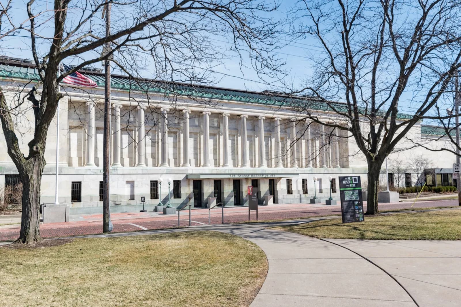 Sunny view of the neoclassical colonnaded facade of the Toledo Museum of Art in Toledo, Ohio, with its tall columns, green roof, and American flag, seen from the lawn and trees in front.
