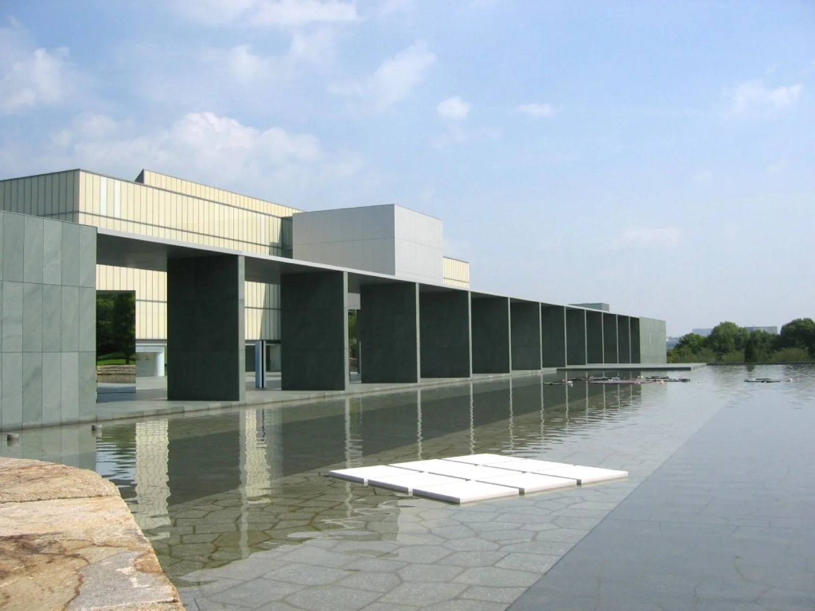 The Toyota Municipal Museum of Art in Toyota, Japan, a modern building with large stone and glass facade, viewed from the water garden on a sunny day.