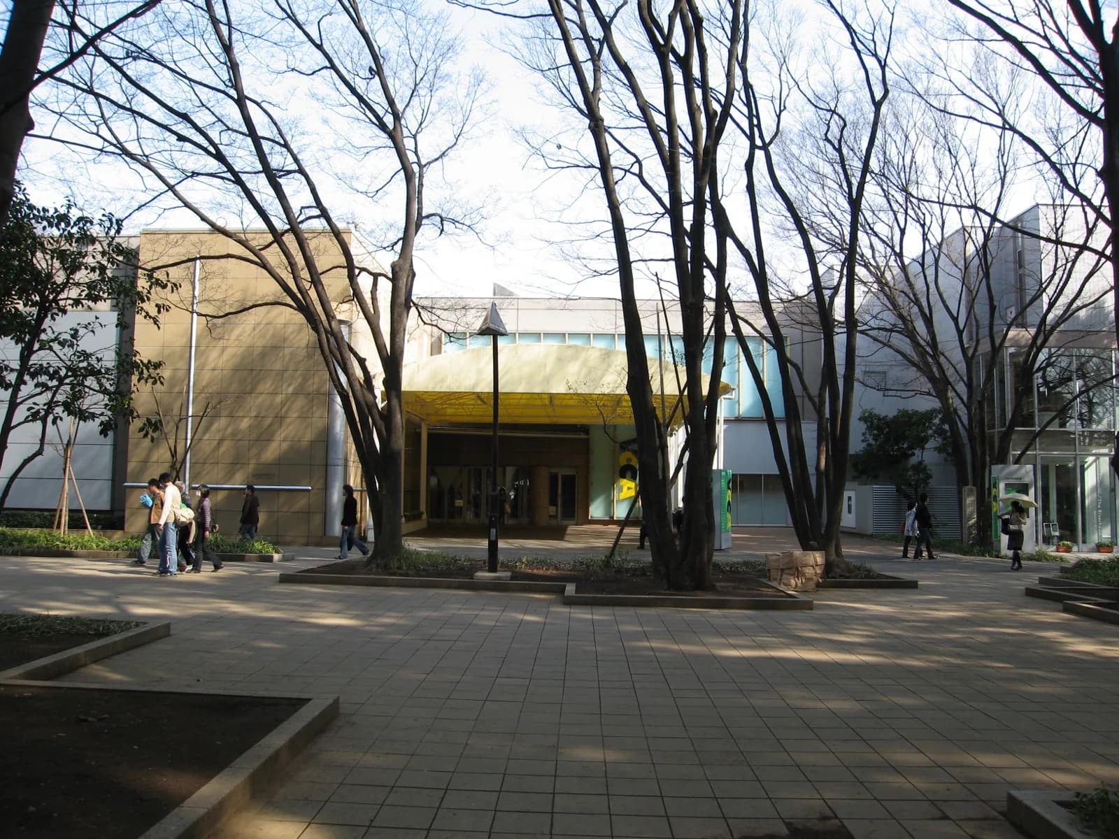 The modern exterior of the Ueno Royal Museum in Ueno Park, Tokyo, featuring a beige tiled facade with a prominent yellow entrance canopy, viewed across a paved plaza with bare trees on a sunny day.