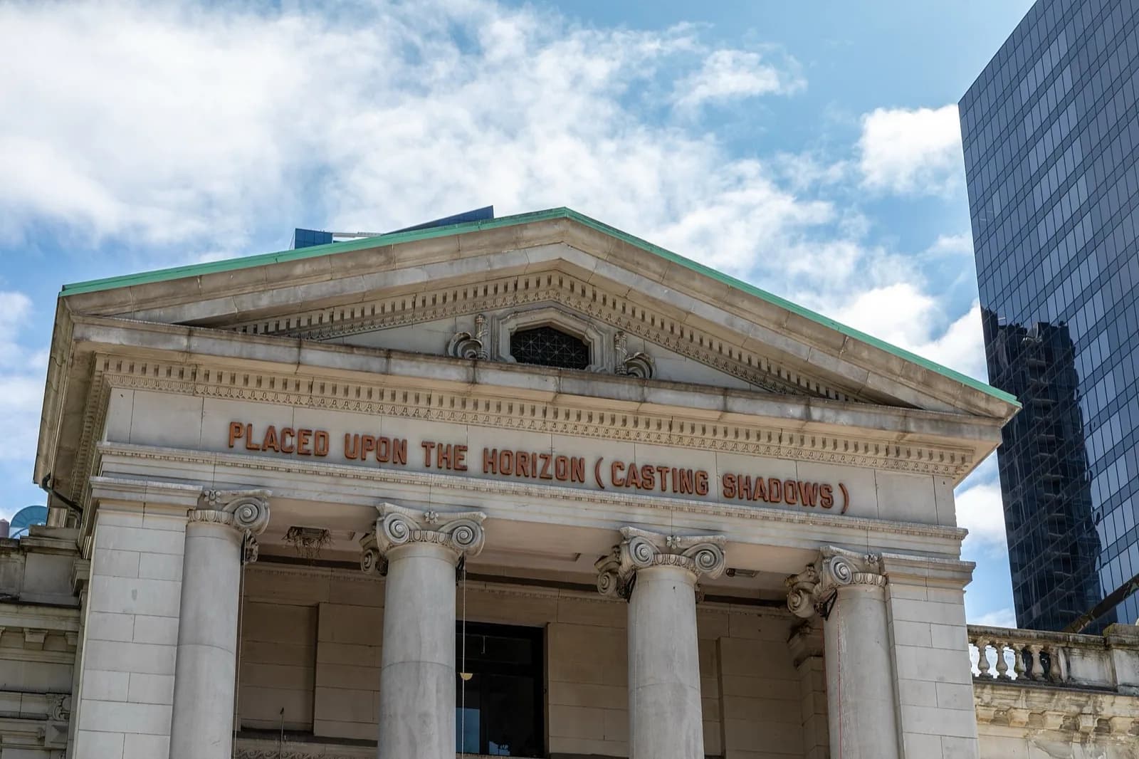Neoclassical stone facade of the Vancouver Art Gallery in Vancouver, Canada, showing the grand portico with tall columns and pediment bearing the inscription 'PLACED UPON THE HORIZON (CASTING SHADOWS)', under a blue sky beside a modern glass skyscraper.