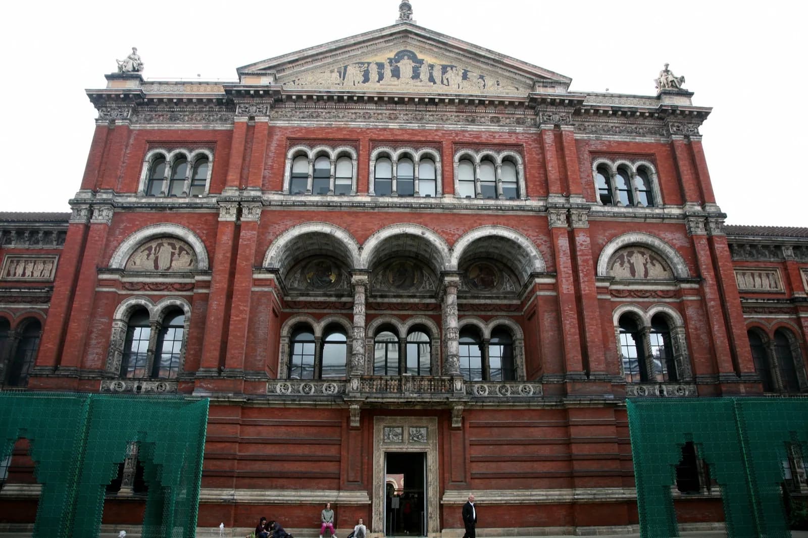 Ornate Victorian red brick and Portland stone facade of the Victoria and Albert Museum along Cromwell Road, London, with arched windows, decorative details, a tower, and foreground garden under a bright sky.