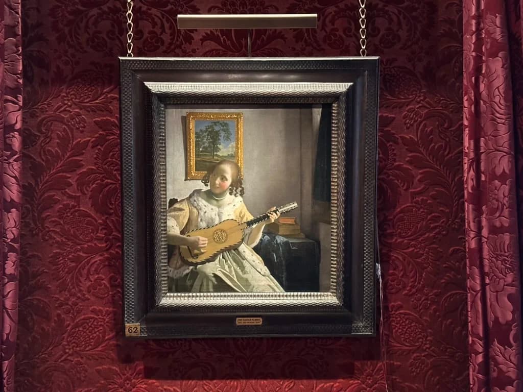 Vermeer's The Guitar Player in a black and silver frame, lit from above, hanging on red damask at Kenwood House, with the gallery number 62 on a label below it.