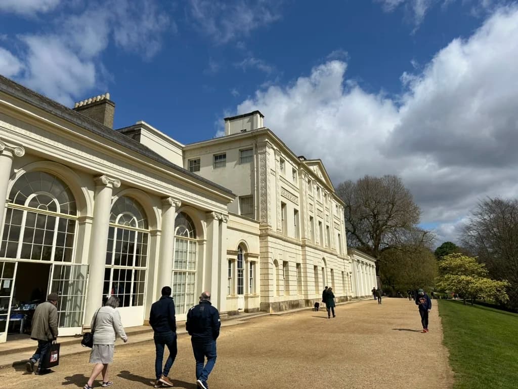 The south side of Kenwood House seen along a gravel path, with the tall arched windows of the Orangery to the left and visitors walking beside the building past the lawns of Hampstead Heath.
