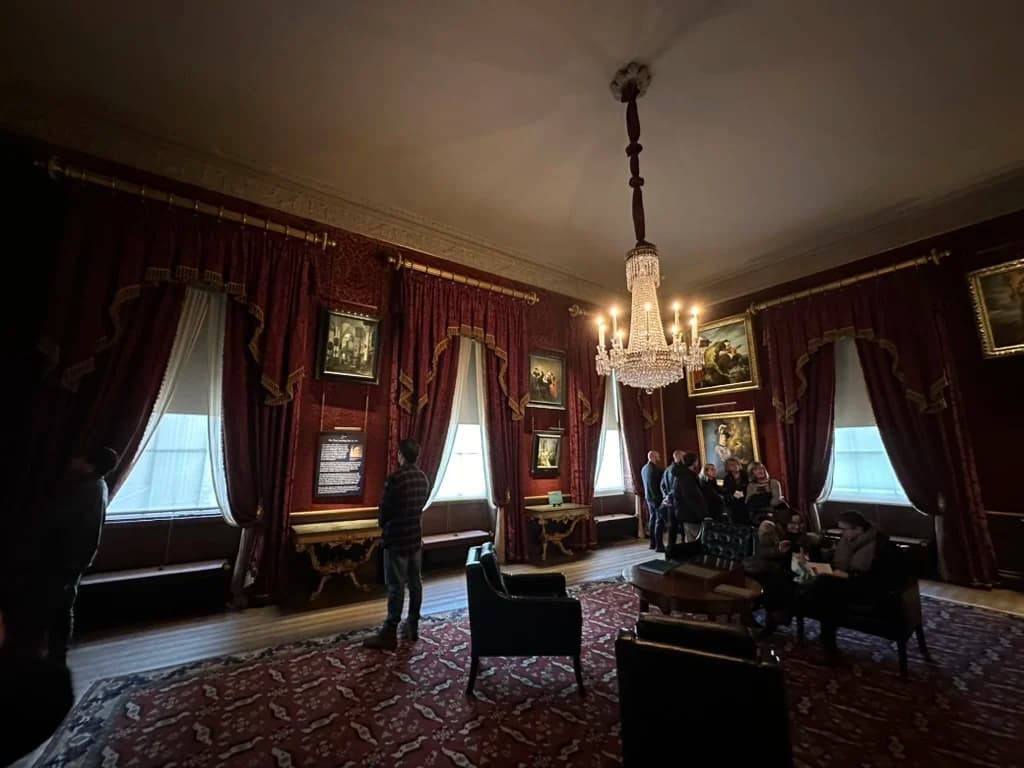 An interior room at Kenwood with deep red damask walls and matching curtains, hung with Old Master paintings in gilt frames and lit by a crystal chandelier, with visitors looking at the pictures.
