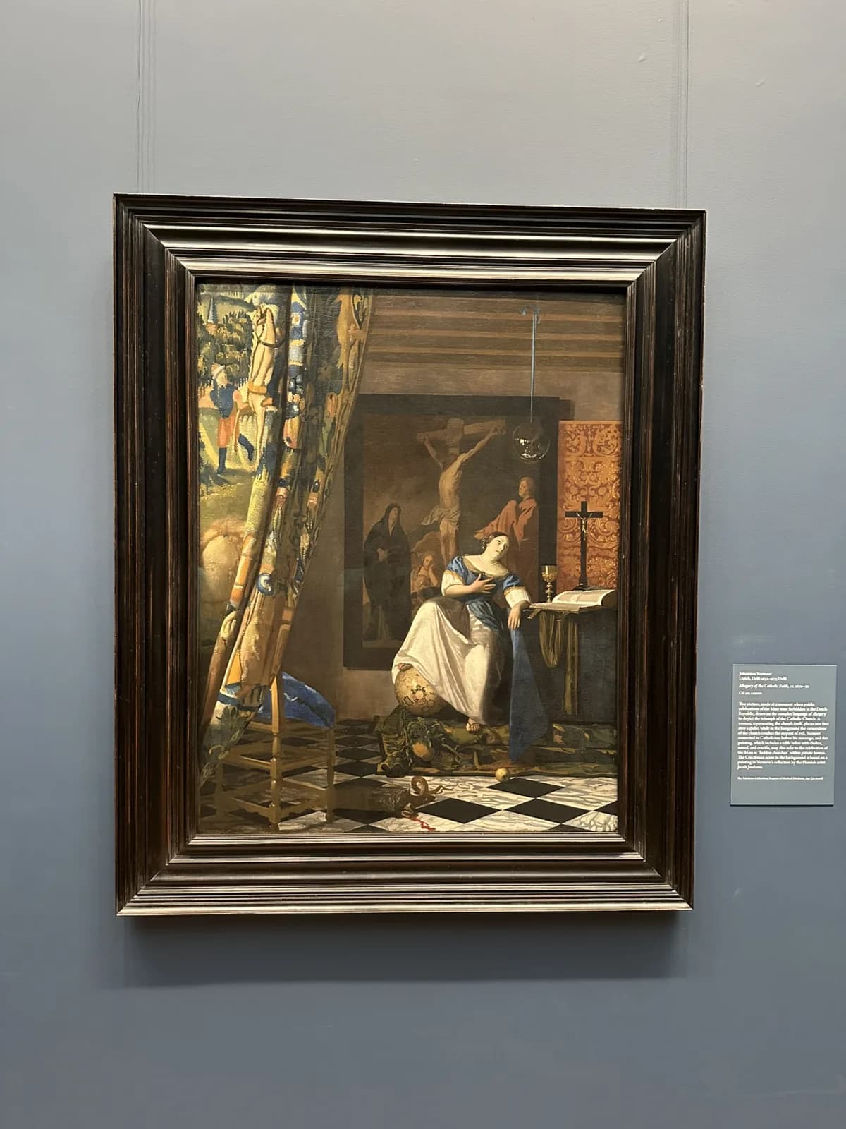 Vermeer's Allegory of Faith hanging in a heavy dark frame on a grey-blue gallery wall at the Met, with its wall label to the right.