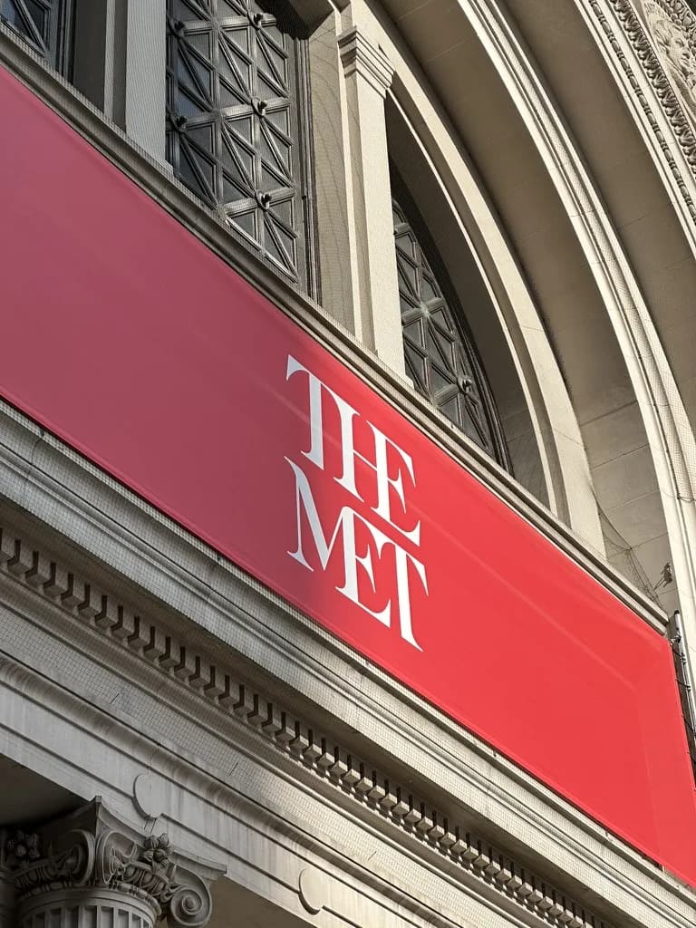 The Met's red banner with the museum's wordmark "THE MET" in white, mounted across the limestone arches of the museum's Beaux-Arts facade on Fifth Avenue.