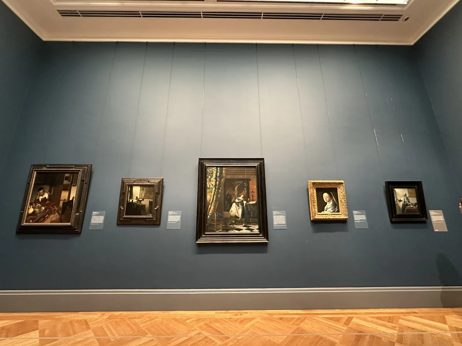 A blue-grey gallery wall in the Met's European paintings rooms hung with five small Dutch Golden Age paintings in dark frames, each accompanied by a wall label, above a herringbone parquet floor.