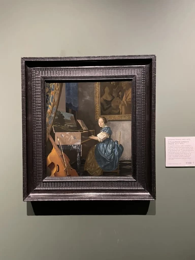Vermeer's A Lady Seated at a Virginal in a dark rippled frame on a sage-green gallery wall at the National Gallery, with its wall label to the right.