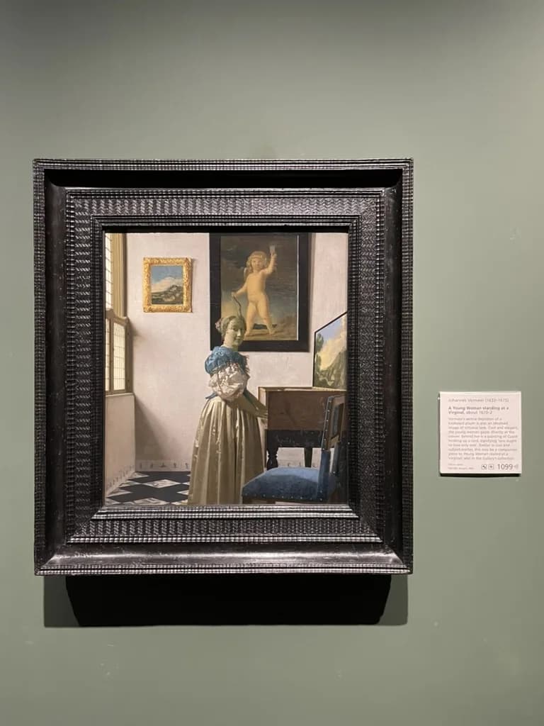 Vermeer's A Lady Standing at a Virginal in a dark rippled frame on a sage-green gallery wall at the National Gallery, with its wall label to the right.