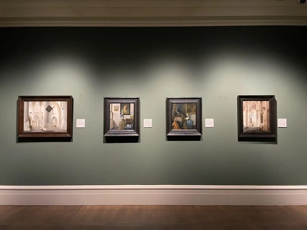 A sage-green gallery wall at the National Gallery hung with four Dutch Golden Age paintings in dark frames, the two central works being Vermeer's A Lady Seated at a Virginal and A Lady Standing at a Virginal, flanked by church interiors, above a wooden floor.